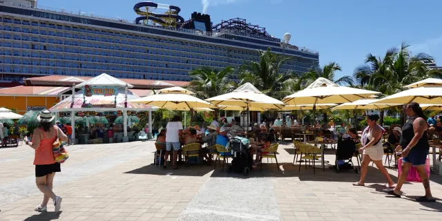 Outdoor seating area with yellow umbrellas at the Coxen Hole cruise port in Roatán, with the Norwegian Escape cruise ship docked behind palm trees.