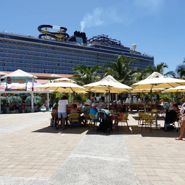 Outdoor seating area with yellow umbrellas at the Coxen Hole cruise port in Roatán, with the Norwegian Escape cruise ship docked behind palm trees.